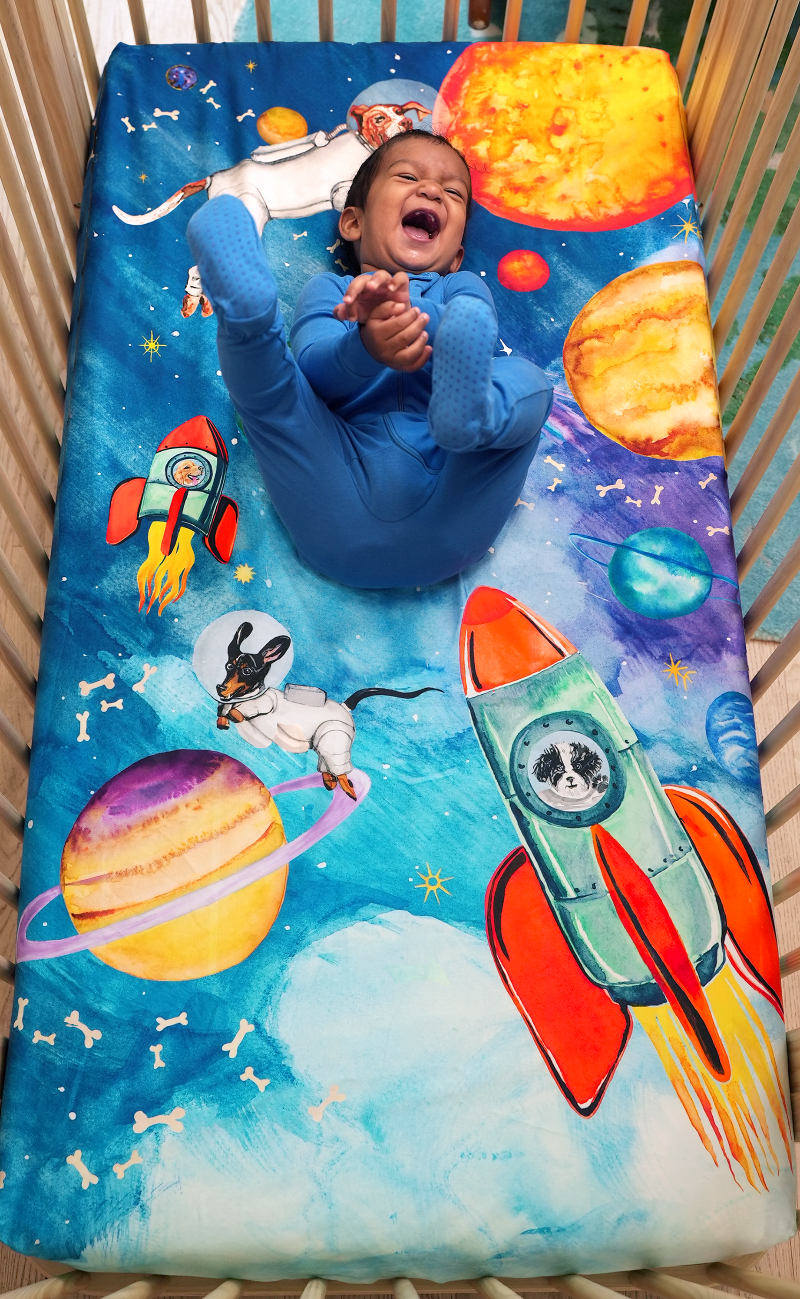 Space themed crib sheet with watercolored designs of dogs dressed as astronauts floating in space and in rocket ships. Design features planets, stars, and floating dog bones. 100% cotton crib sheet, fits US standard size cribs.