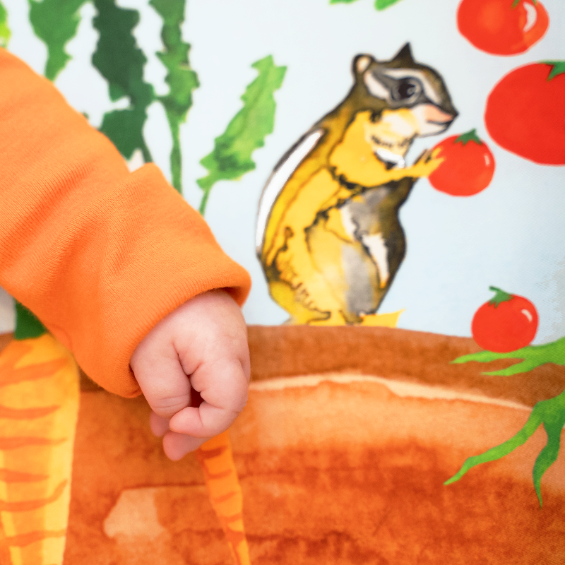 Detail shot of a baby's hand laying on top of a vegetable garden themed sheet featuring a watercolor painting of a chipmunk snacking on a tomato.