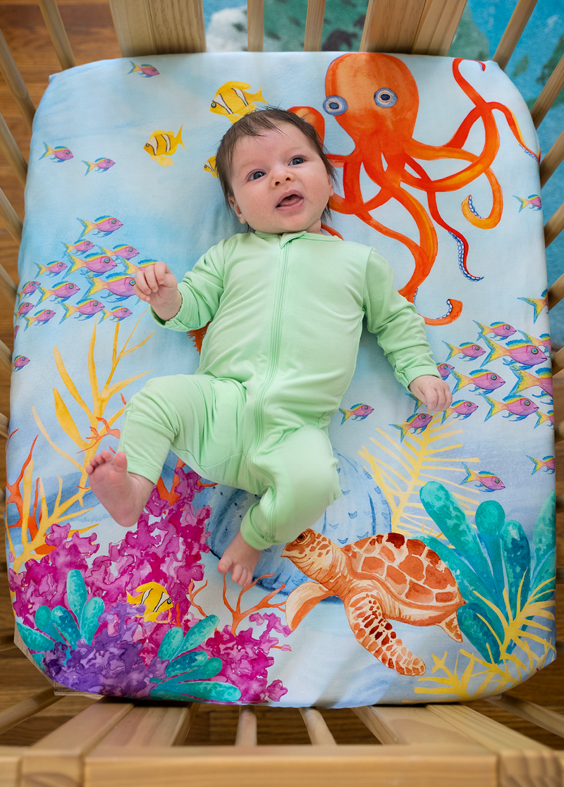 Newborn baby laying on top of a sea themed nestig compatible mini crib sheet featuring a bright red-orange octopus, pink and yellow fish, colorful coral plants, and a sea turtle