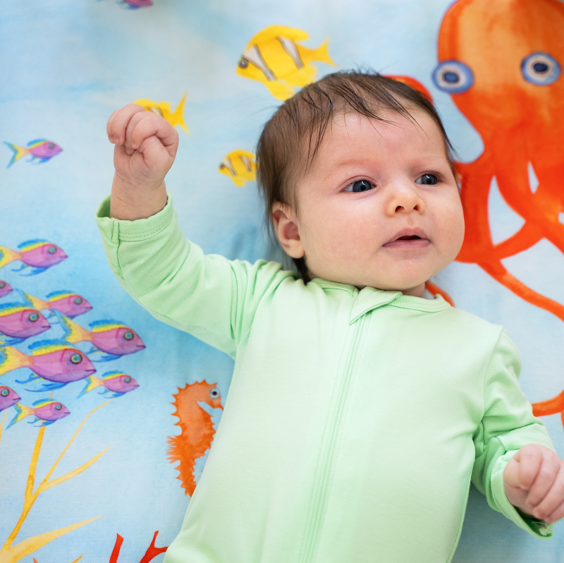 Newborn baby laying on top of a sea themed nestig compatible mini crib sheet featuring a bright red-orange octopus, pink and yellow fish, colorful coral plants, and a sea turtle