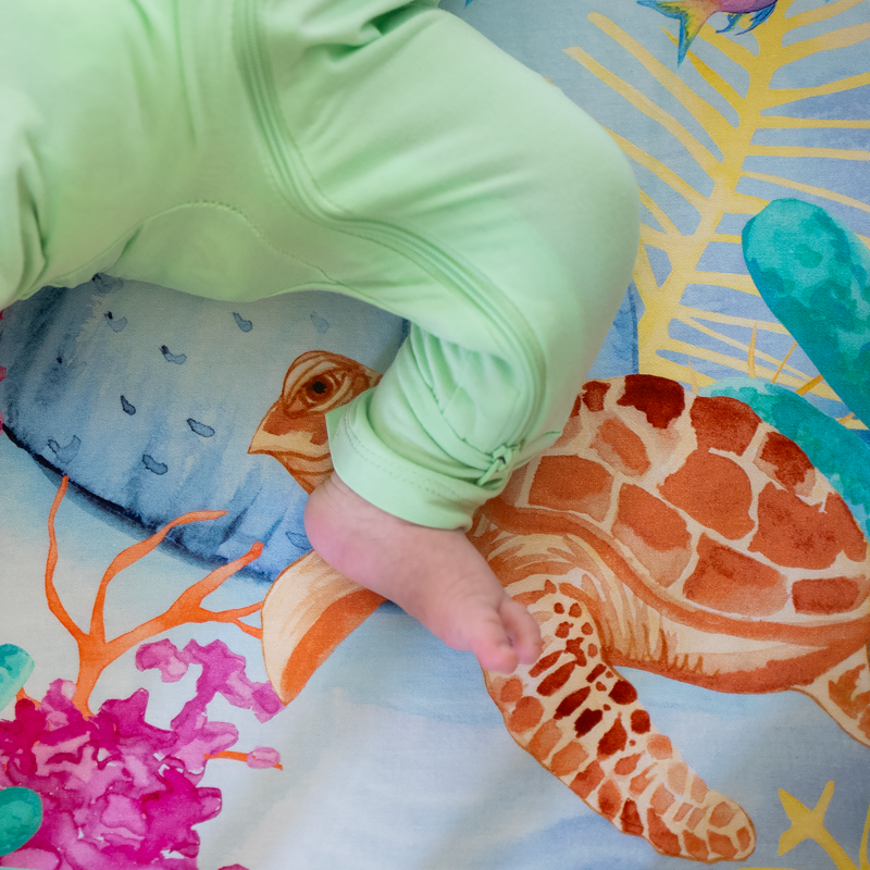 Newborn baby laying on top of a sea themed nestig compatible mini crib sheet featuring a bright red-orange octopus, pink and yellow fish, colorful coral plants, and a sea turtle