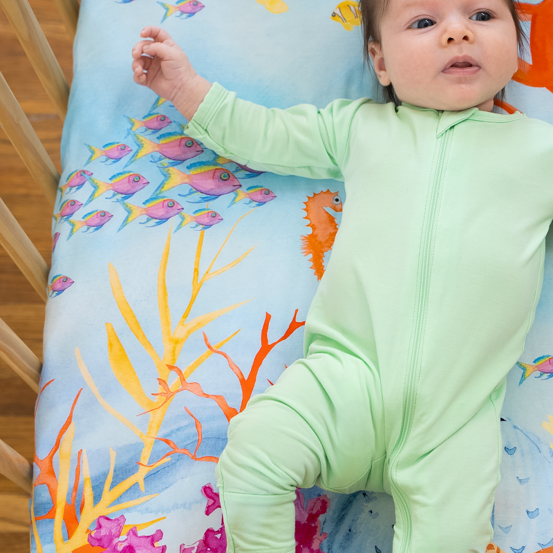 Newborn baby laying on top of a sea themed nestig compatible mini crib sheet featuring a bright red-orange octopus, pink and yellow fish, colorful coral plants, and a sea turtle