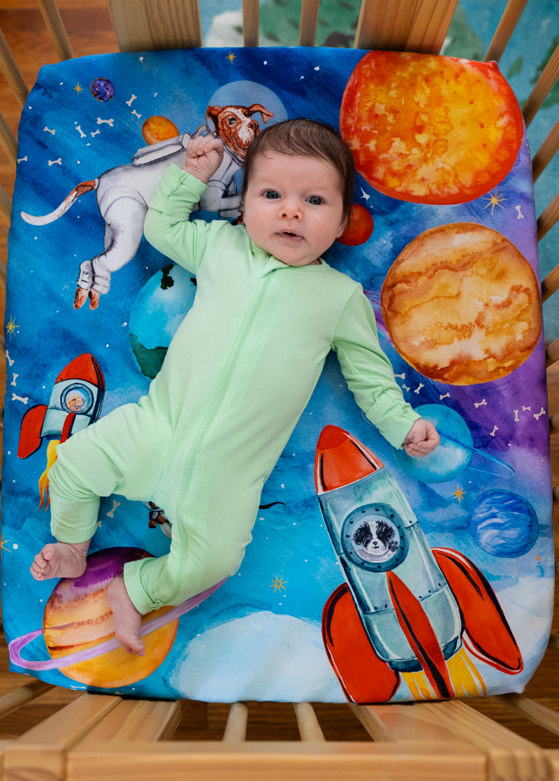 Newborn baby laying on top of an outer-space themed nestig compatible mini crib sheet featuring a dogs dressed as astronauts, flying in rockets and surrounded by the solar system, shooting starts, and floating dog bones.
