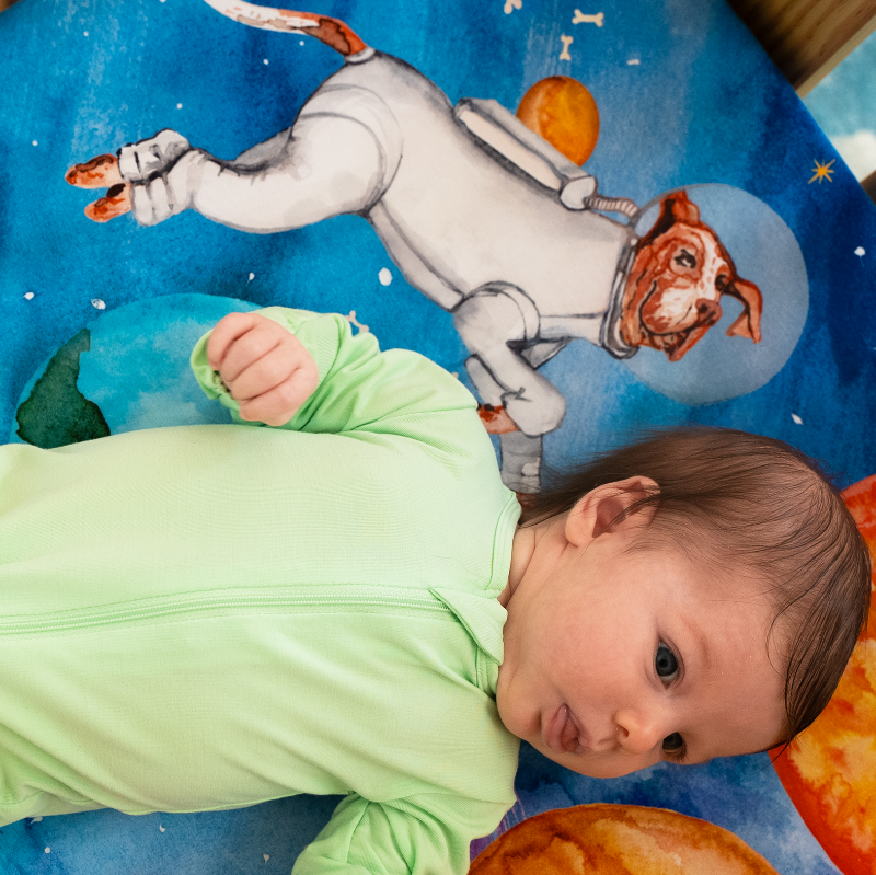 a close up shot of a baby's face laying on top of a nestig mini crib sheet featuring a hound dressed as an astronaut