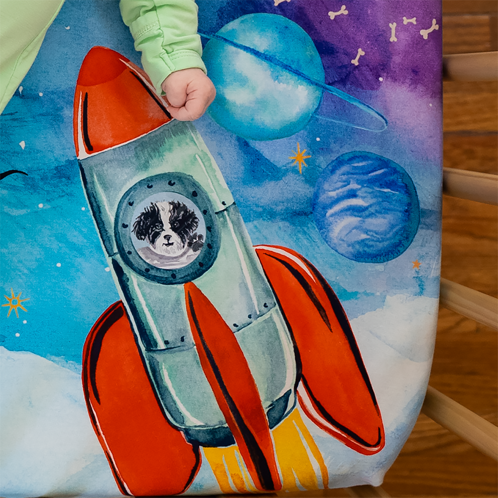 Close up shot of mini sheet featuring a baby's fist next to an illustration of a black and white poodle riding a rocket ship.