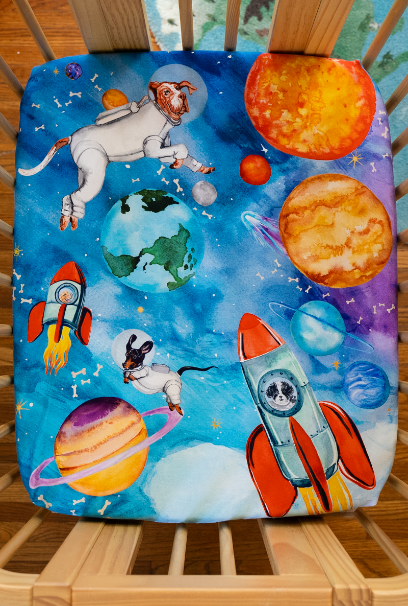 nestig compatible mini crib sheet featuring a hand water colored design of astronaut dogs floating in outer-space surrounded by the solar system, floating bones, and stars.
