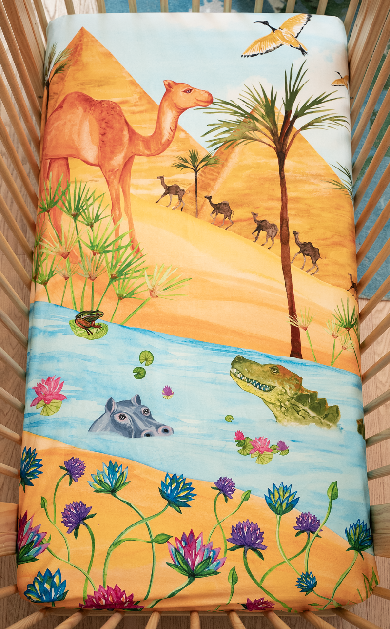 US standard size crib sheet, Naptime on the Nile. Watercolor design featuring the Egyptian pyramids, Nile river, palm trees, sand dunes, camels, lotus flowers, reeds, a crocodile, hippo, lily pads, and a frog.