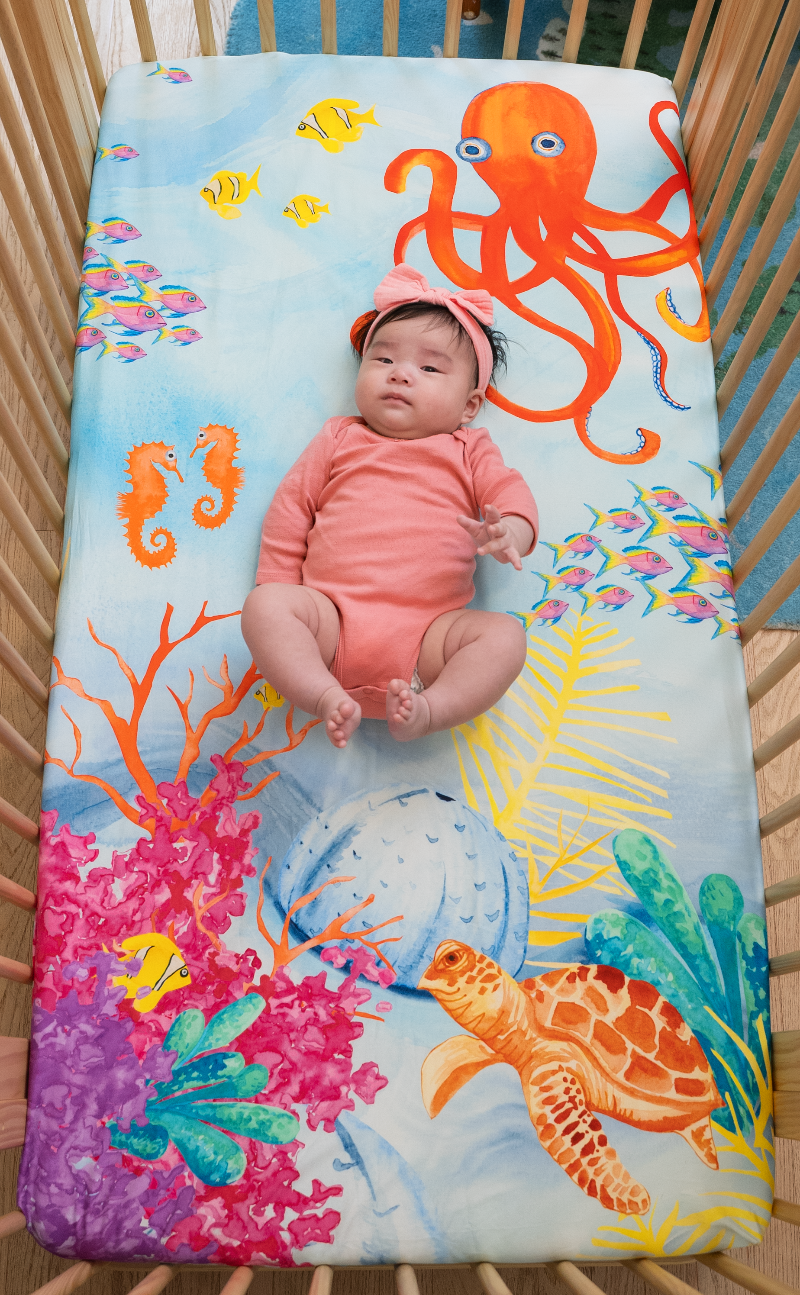 Ocean theme crib sheet, Sea You In My Dreams. Watercolor design features sea creatures such as a big red-orange octopus, tropical fish, vibrant colored coral, and a friendly sea turtle. Fits US standard mattresses.