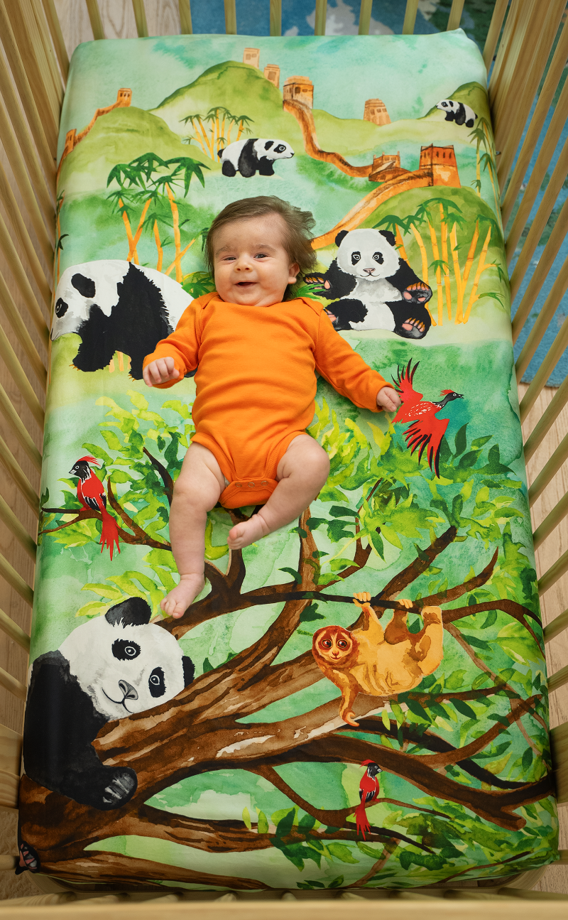 US standard size crib sheet, Great Cuddle of Pandas that features hand watercolored design of pandas lounging on forest trees, eating bamboo, a sloth hanging on a tree with red birds flying. Lush, green forest scene with mountains and The Great Wall of China in the distance.
