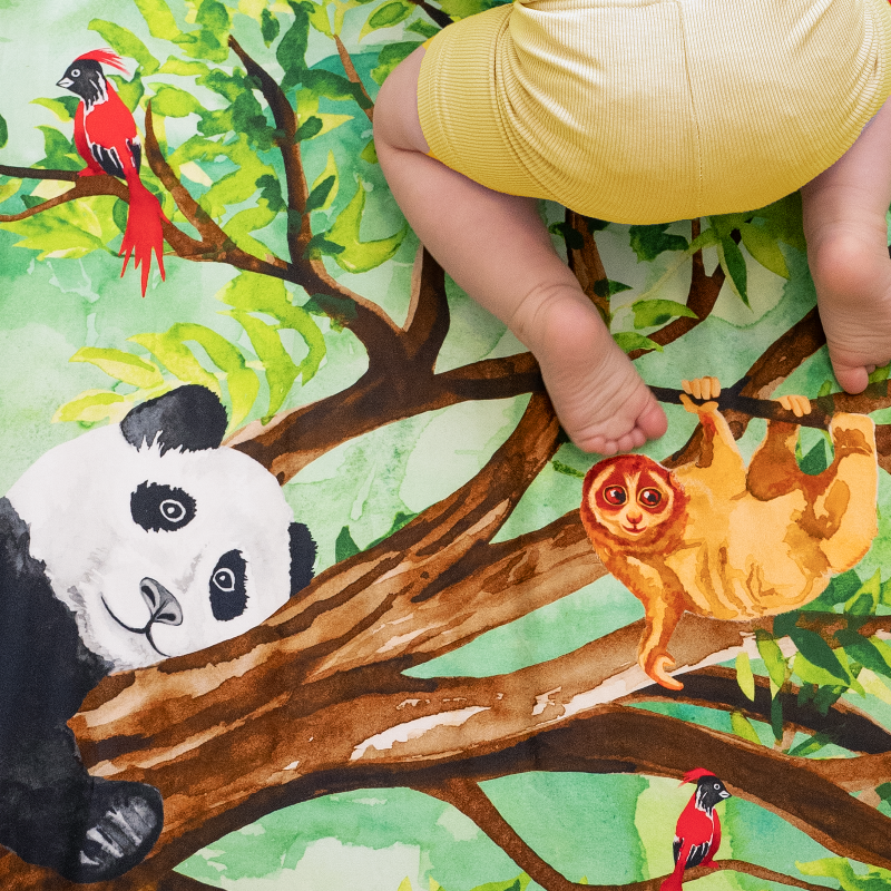 Detail of Great Cuddle of Pandas crib sheet. Watercolor depiction of a panda resting in a lush green tree, next to a swinging sloth and perched red birds.