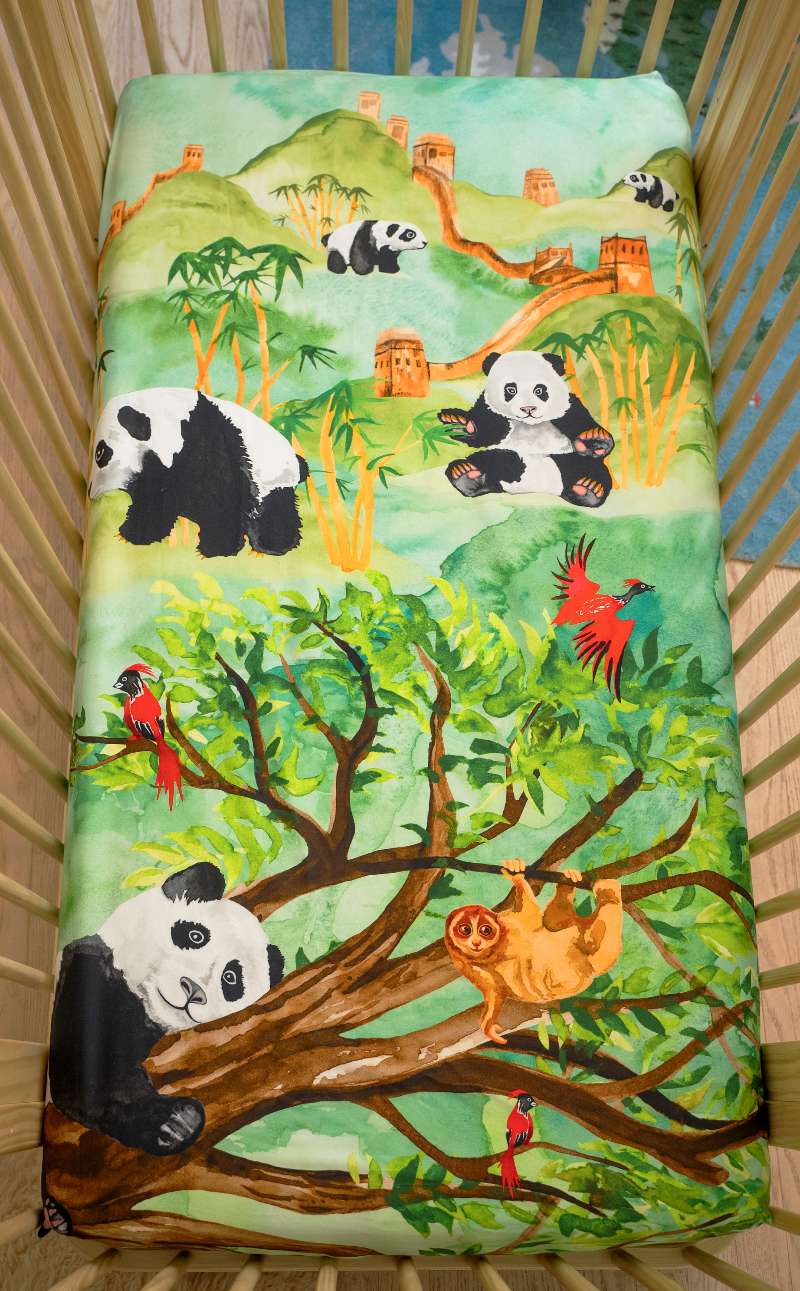 US standard size crib sheet, Great Cuddle of Pandas that features hand watercolored design of pandas lounging on forest trees, eating bamboo, a sloth hanging on a tree with red birds flying. Lush, green forest scene with mountains and The Great Wall of China in the distance.