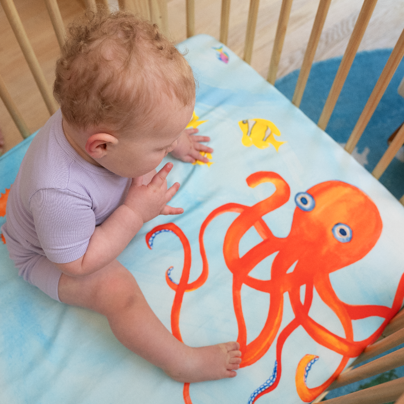 Detail of Sea You In My Dreams crib sheet showing a baby sitting on top of the ocean crib sheet looking down at the watercolor depiction of an orange octopus. 100% cotton crib sheet.