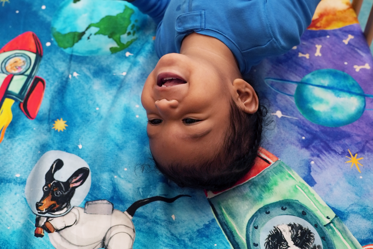 toddler smiling on top of a space theme crib sheet featuring watercolored designs of dogs dressed as astronauts floating in space and in rocket ships. Design features planets, stars, and floating dog bones. 100% cotton crib sheet, fits US standard size cribs.