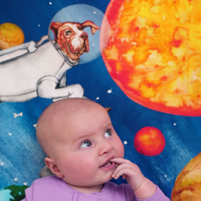 baby on top of a space theme crib sheet featuring watercolored designs of a dog dressed as an astronauts floating in space with planets and stars. 100% cotton crib sheet, fits US standard size cribs.