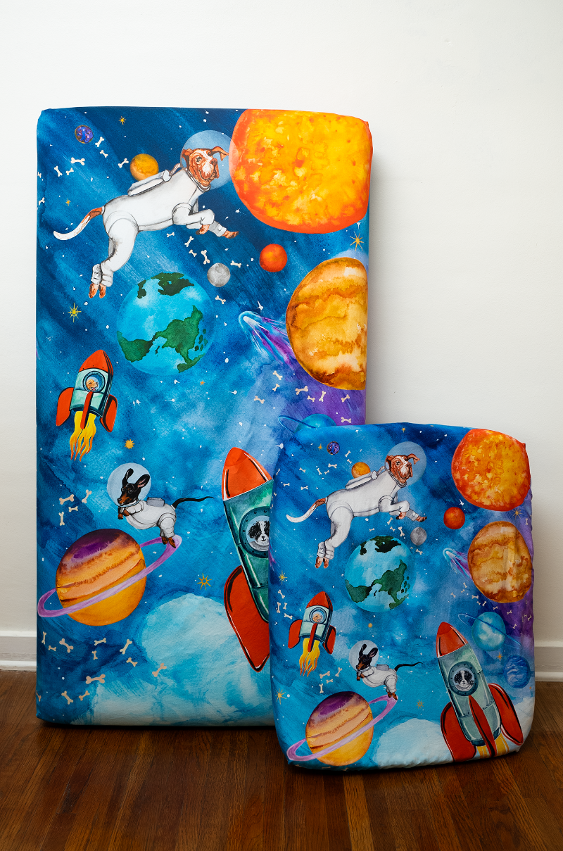A full size crib sheet next to a nestig mini compatible crib sheet both in the same outer-space design featuring a dogs dressed as astronauts, flying in rockets and surrounded by the solar system, shooting starts, and floating dog bones.