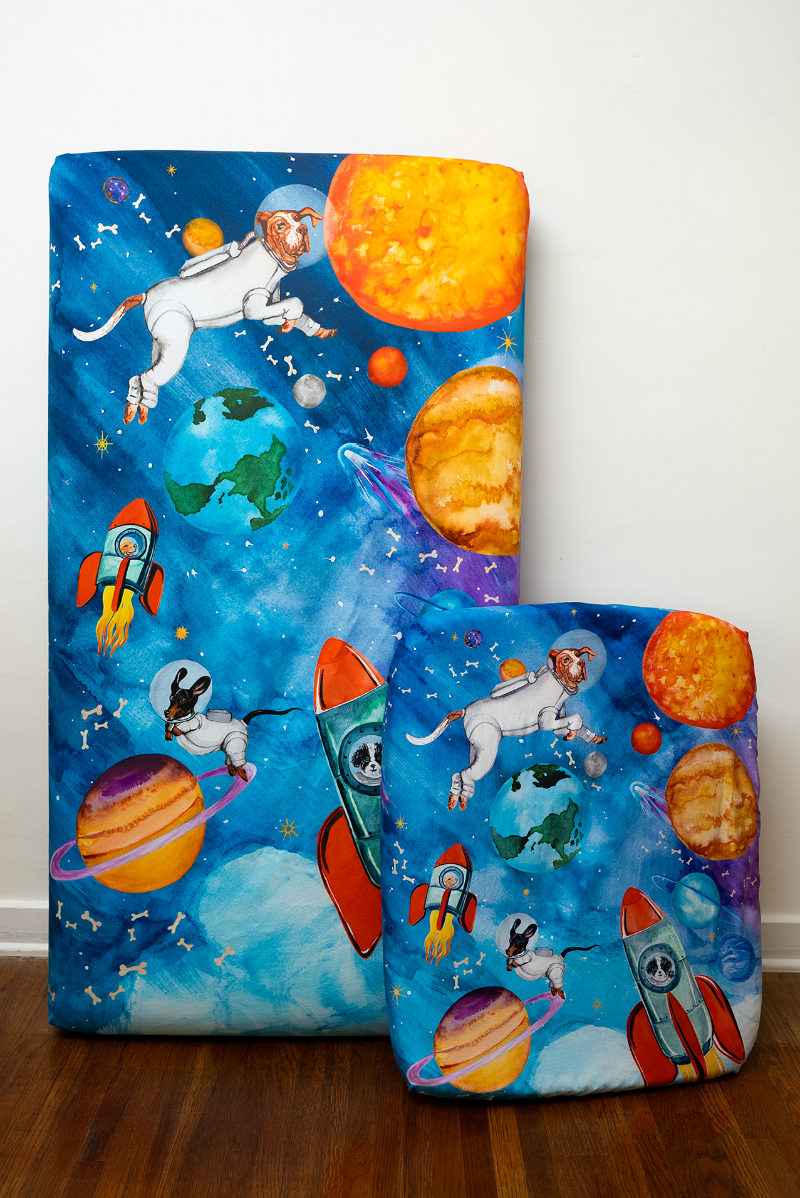 A full size crib sheet next to a nestig mini compatible crib sheet both in the same outer-space design featuring a dogs dressed as astronauts, flying in rockets and surrounded by the solar system, shooting starts, and floating dog bones.