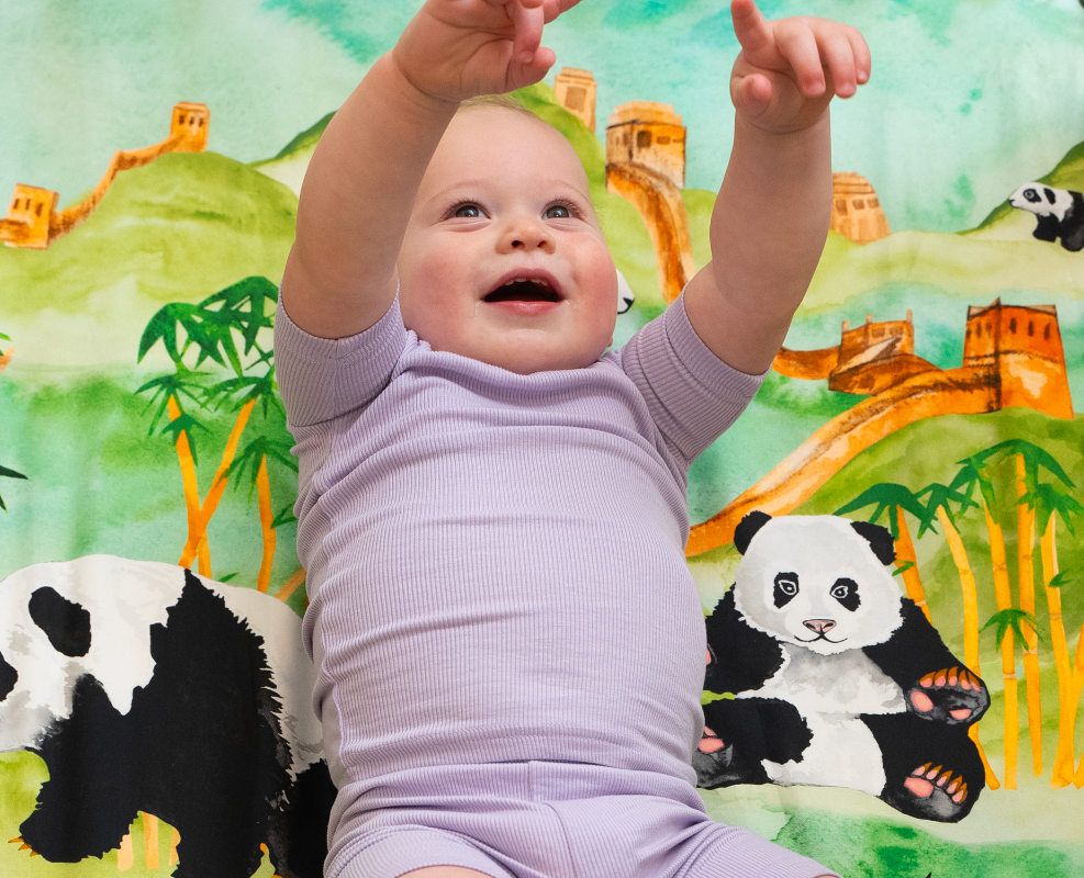happy baby laying on top of a panda sheet featuring water color paintings of pandas, bambo, lush green forest and mountains, and The Great Wall of China in the distance.