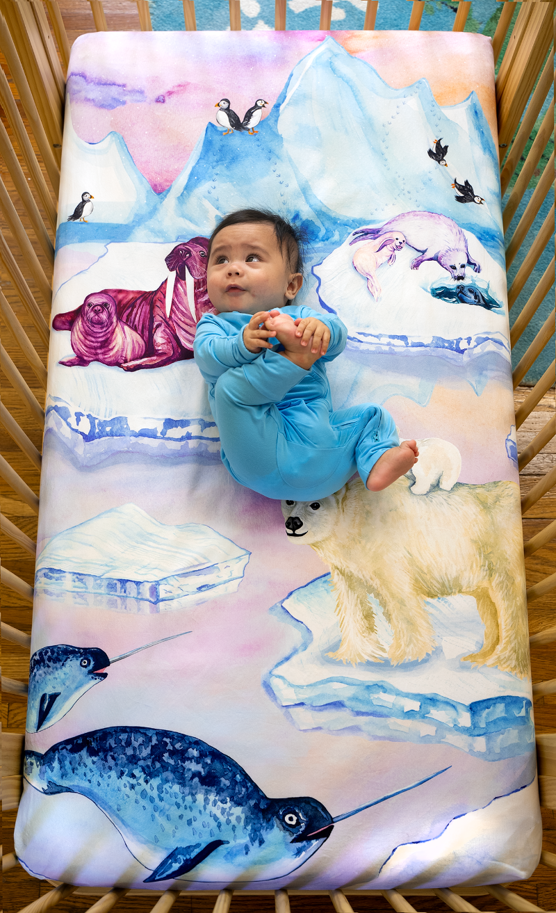 happy baby laying on top of an arctic themed crib sheet featuring seals, walruses, polar bears, narwhals, and puffins in a snowy, icy scene