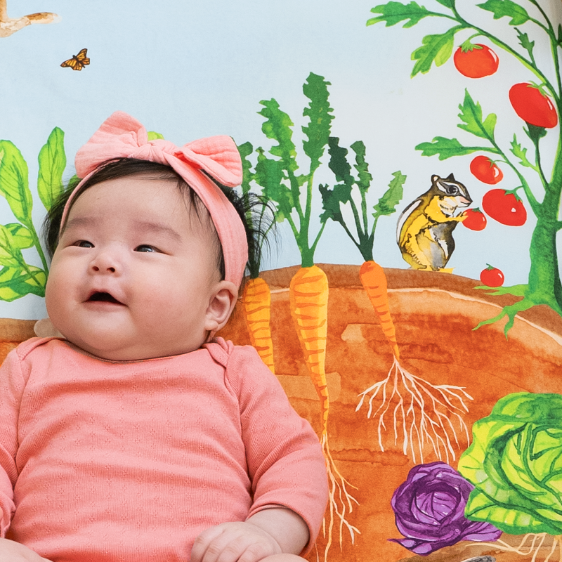 Curious baby laying on top of a vegetable garden themed crib sheet featuring watercolor paintings of a chipmunk munching on a tomato plant, carrots and cabbages growing underground, and butterflies flying in the sky.