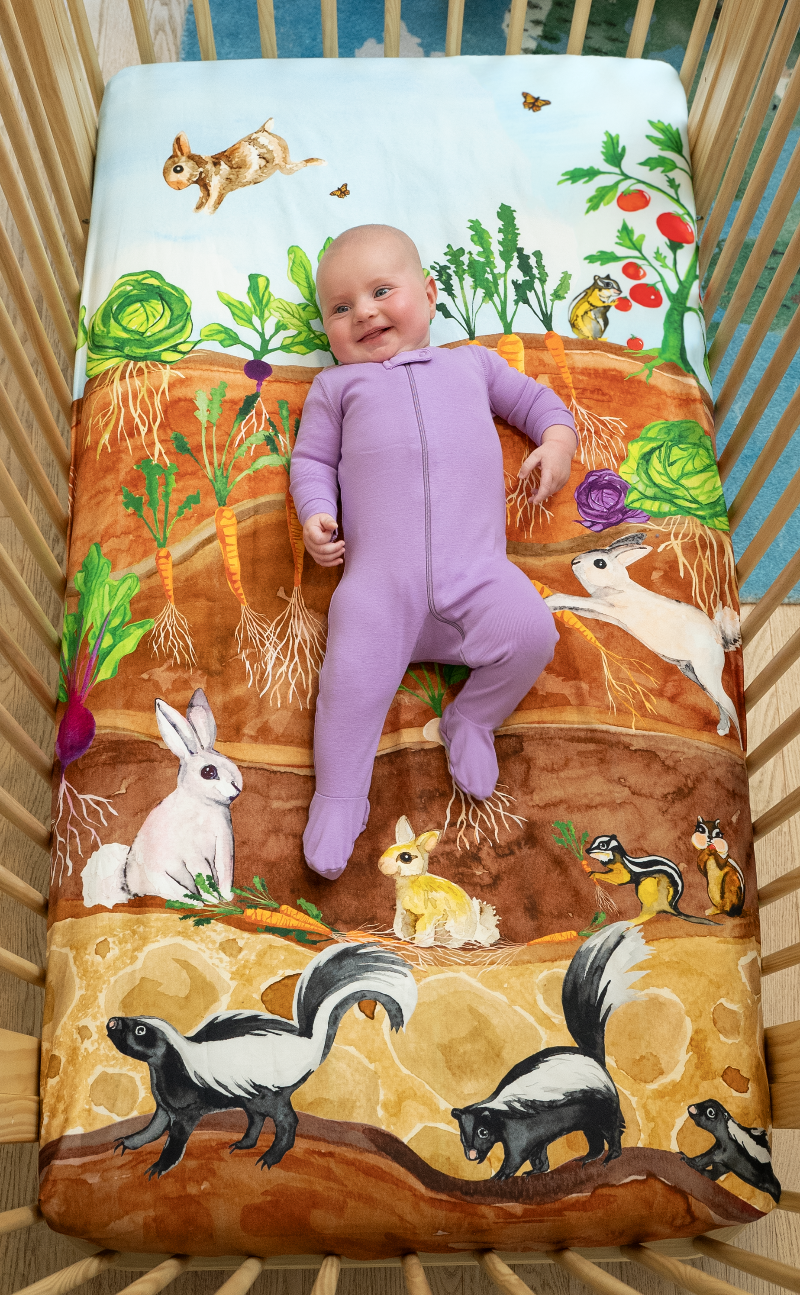 US Standard size crib sheet, Garden Bed Burrow. Watercolor design featuring a vegetable garden of cabbages, radishes, carrots, and tomatoes. Bunny rabbits, butterflies, chipmunks, and skunks inhabit this playful scene.