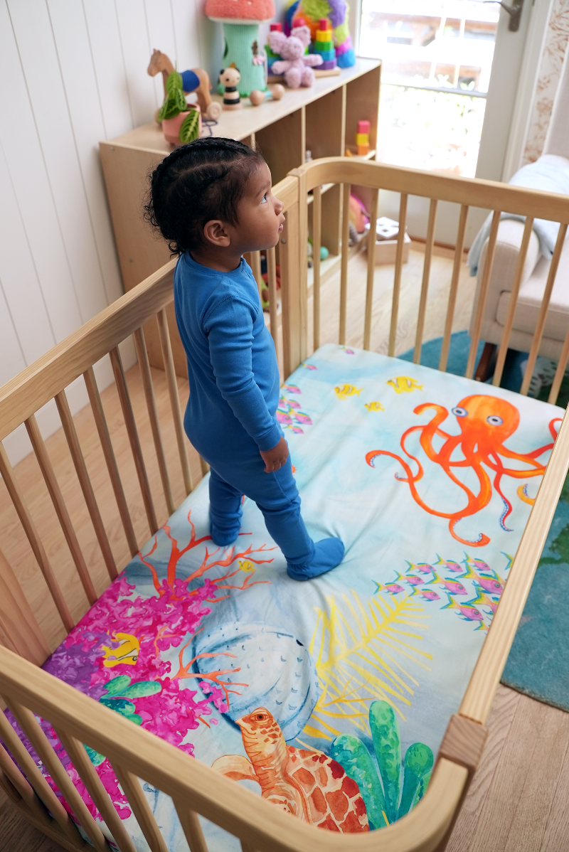 toddler standing on top of a sea themed crib sheet featuring a bright red-orange octopus, yellow and pink tropical fish, colorful coral, and a friendly sea turtle.