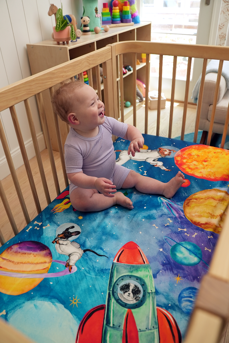 Baby laughing and sitting on top of an outer-space themed crib sheet featuring dogs dressed as astronauts floating amongst the solar system.